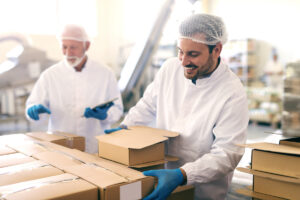 man relocating boxes with cookies in food factory. In background his boss counting boxes and holding tablet | Sirocco Food Safety Consulting